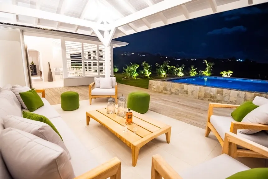 Night view of a luxurious open-air patio with white and green seating, wooden table, and a lit swimming pool in the background.