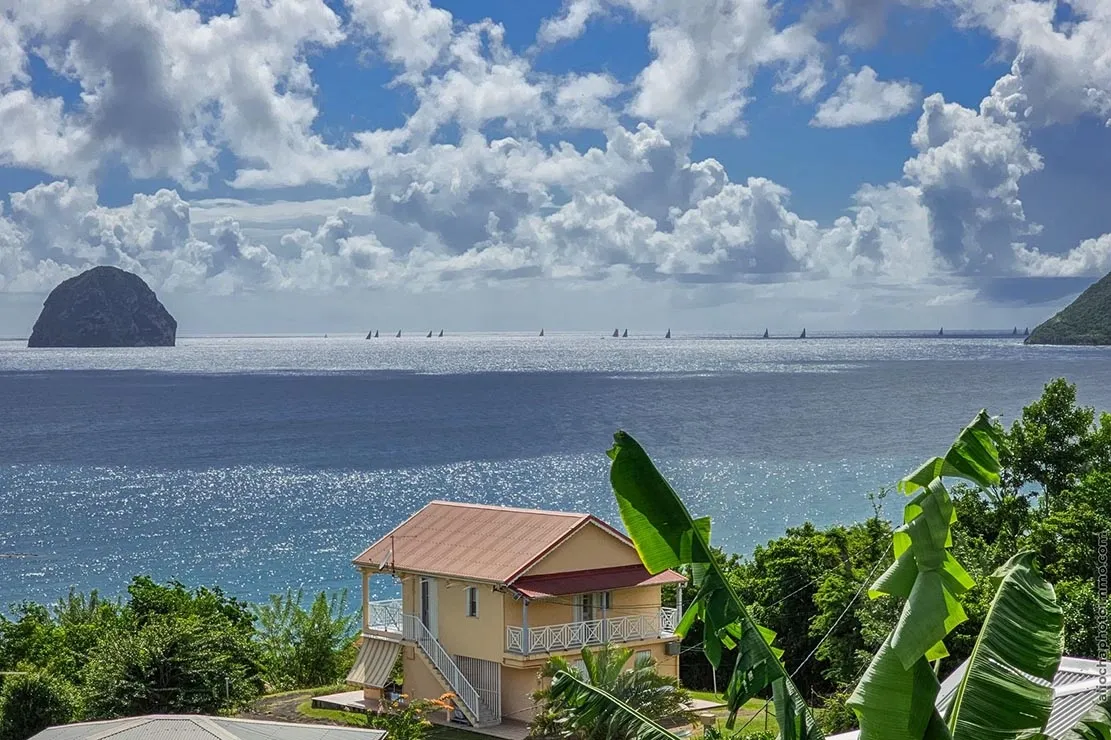 Yellow villa with red roof overlooking the sparkling sea, lush greenery, and a rocky island in Le Diamant, Martinique.