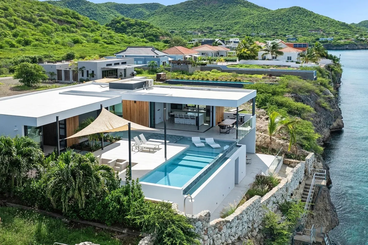 Modern Caribbean villa with infinity pool overlooking the sea and lush green hills in the background.