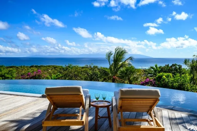 Two lounge chairs by an infinity pool overlooking lush greenery and the ocean under a blue sky in Guadeloupe.